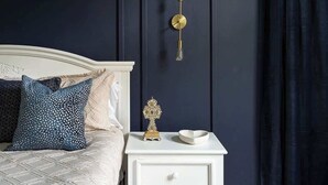 A bedroom with a white bed, dark blue walls, a white nightstand with a decorative item, and a wall-mounted light fixture.