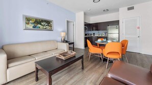 Een woonkamer met een bank, een salontafel en oranje stoelen. Er hangt een schilderij aan de muur en er is een keuken op de achtergrond.
