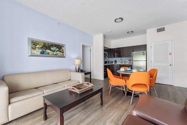 Een woonkamer met een bank, een salontafel en oranje stoelen. Er hangt een schilderij aan de muur en er is een keuken op de achtergrond.