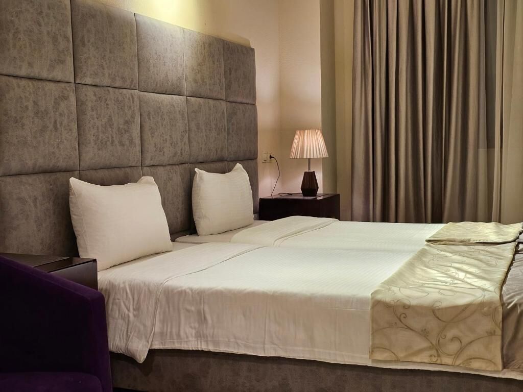 Ett hotellrum med en stor säng, ett vadderat sänggavel, en sänglampa och utsikt över gardinerna.