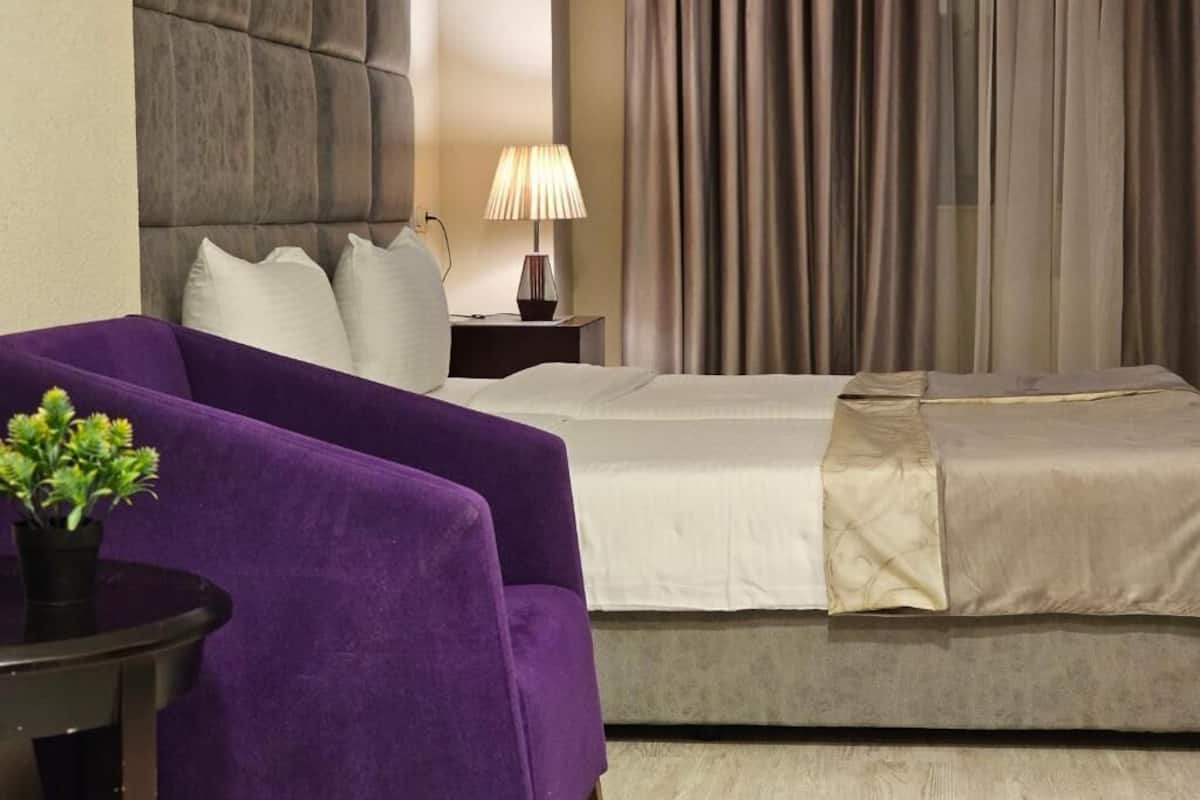 A hotel room with a bed, a nightstand with a lamp, a purple armchair, and a small table with a plant.