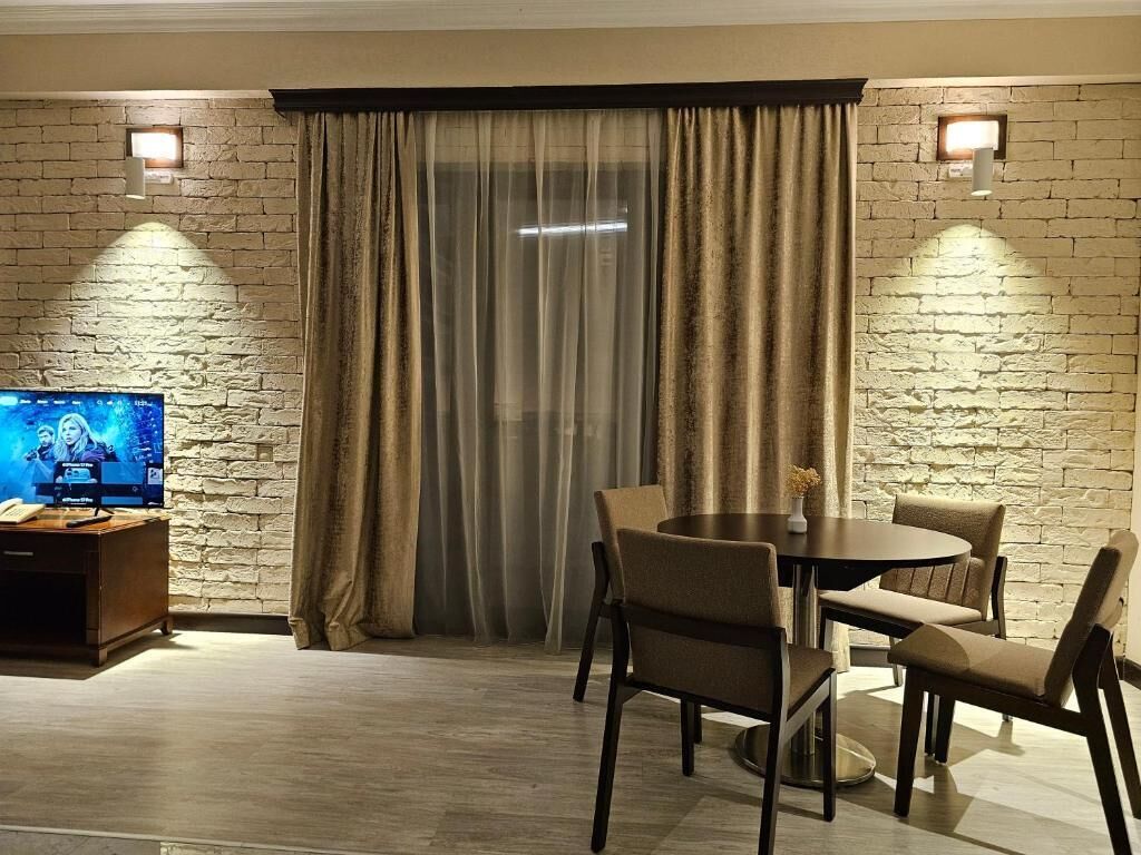 A room with a brick wall, a TV on a wooden stand, a dining table with chairs, and a large window with curtains.