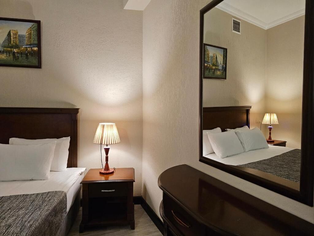 A hotel room with a bed, bedside table with a lamp, a dresser, and a large mirror reflecting the room.