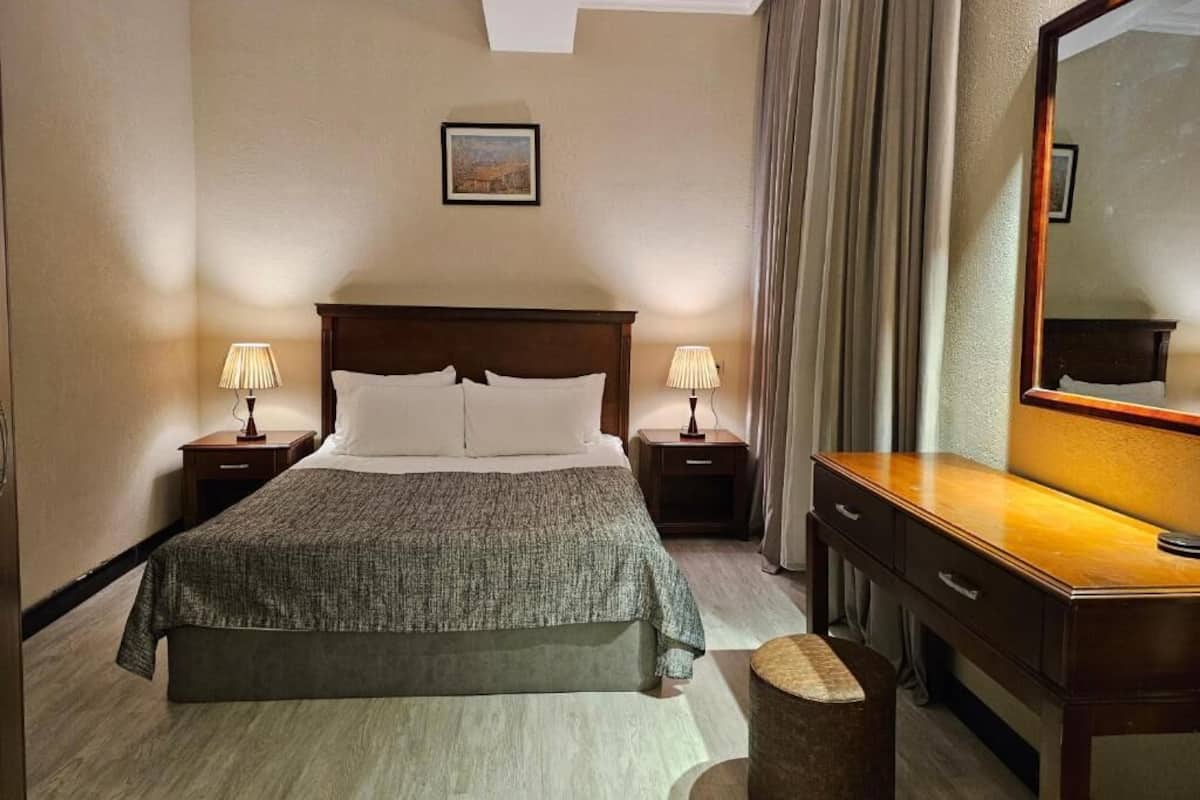 A hotel room with a large bed, two bedside tables with lamps, a dresser, a mirror, and a painting on the wall.