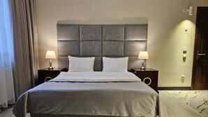 A large bed with a grey headboard, two bedside tables with lamps, and a door to the right. - Crown Suites Hotel (Baku)