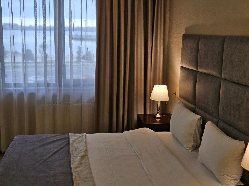 Habitación de hotel con una cama grande, una mesita de noche con lámpara y vistas a una masa de agua a través de cortinas translúcidas.