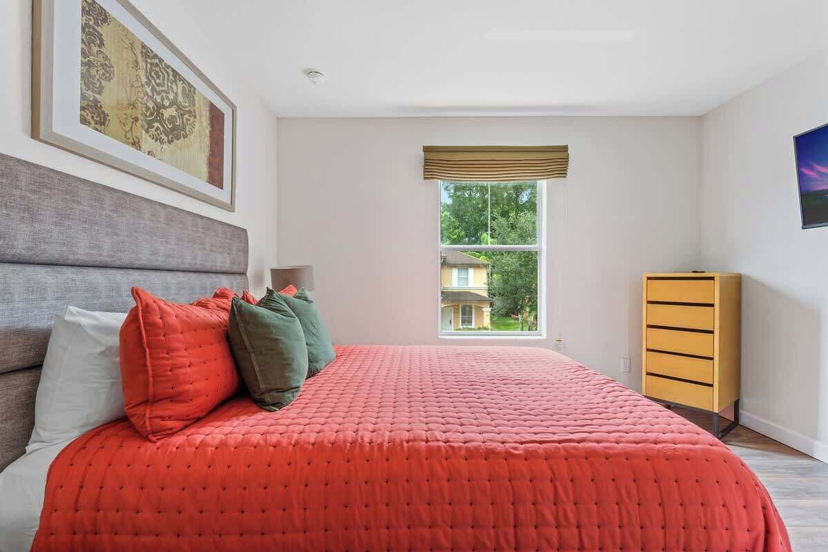 Quarto com cama grande, penteadeira, janela com vista para uma casa e quadro na parede.