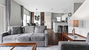 Ein modernes Wohnzimmer mit einem grauen Sofa, einem Couchtisch und einer Lampe. Im Hintergrund ist eine Küche mit einer Theke und Hockern zu sehen.