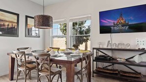 A dining area with a wooden table set for four, surrounded by wicker chairs. A television mounted on the wall displays the Disney logo.