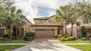 A two-story house with a tiled roof, a garage, and a front yard with palm trees and shrubs. - Pet Friendly Amazing 5 BR sleeps 10 (Kissimmee)