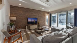 A modern living room with a flat-screen TV, a sofa, a coffee table, and a large window offering a view of a pool.