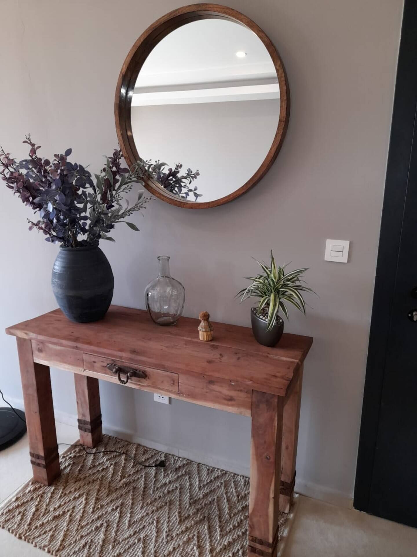 Una consola de madera con un espejo encima, una planta en maceta, un florero y un objeto decorativo pequeño.
