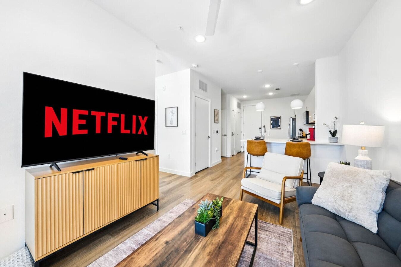 A modern living room with a large flat-screen TV displaying the Netflix logo, a wooden entertainment unit, a grey sofa, a coffee table with a plant, and a kitchen area in the background.