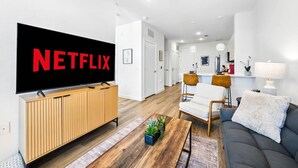 A modern living room with a large flat-screen TV displaying the Netflix logo, a wooden entertainment unit, a grey sofa, a coffee table with a plant, and a kitchen area in the background. - New Modern 1BD/1BA~Pet Love Loft (Austin)