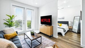 A modern apartment with a living area featuring a sofa, coffee table, and a TV displaying 'NETFLIX'. There is a bedroom with a bed and bedside table. - New Modern 1BD/1BA~Pet Love Loft (Austin)