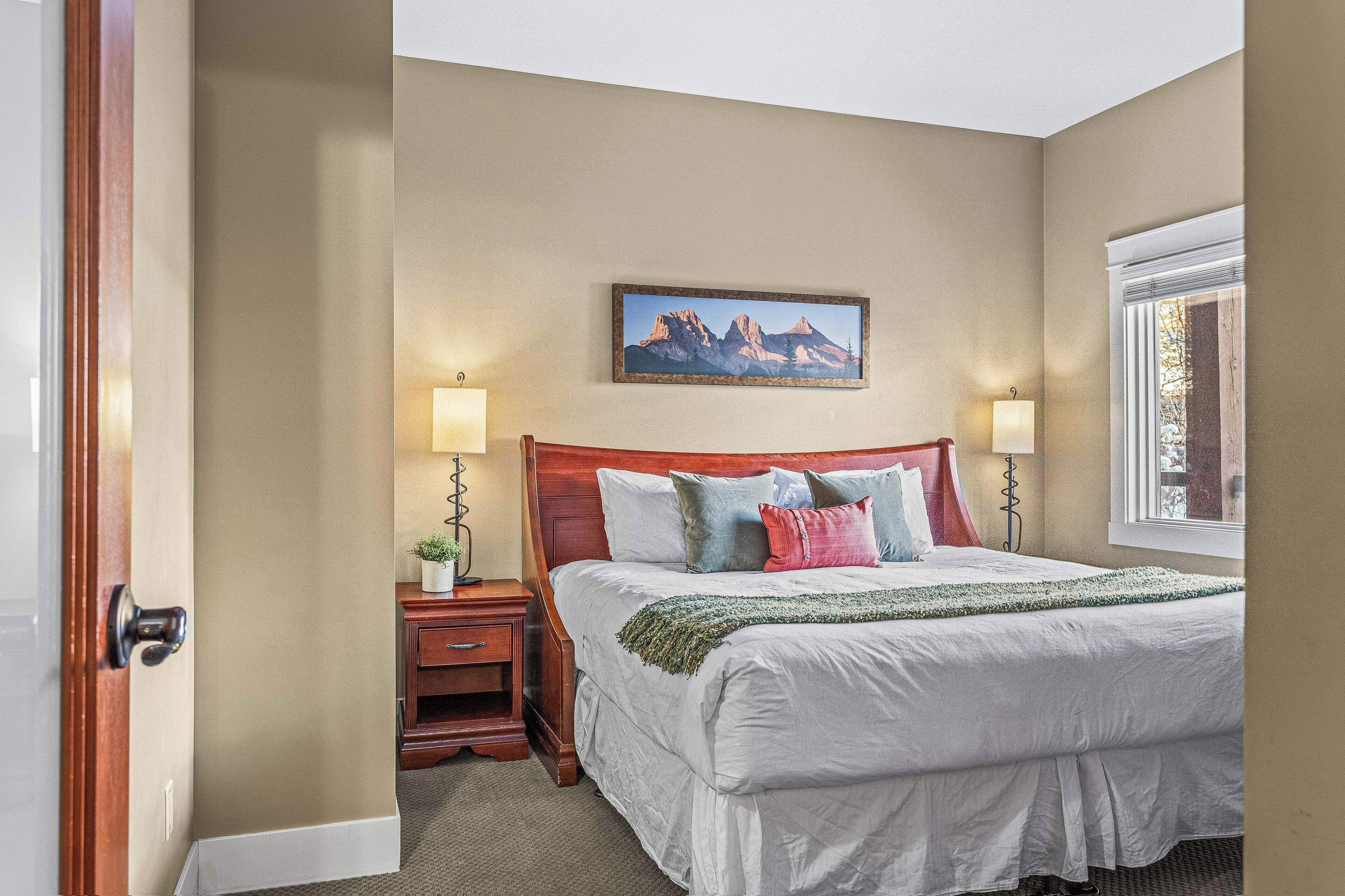 A bedroom with a large bed, two bedside tables, a nightstand, a painting on the wall, and a window with curtains.