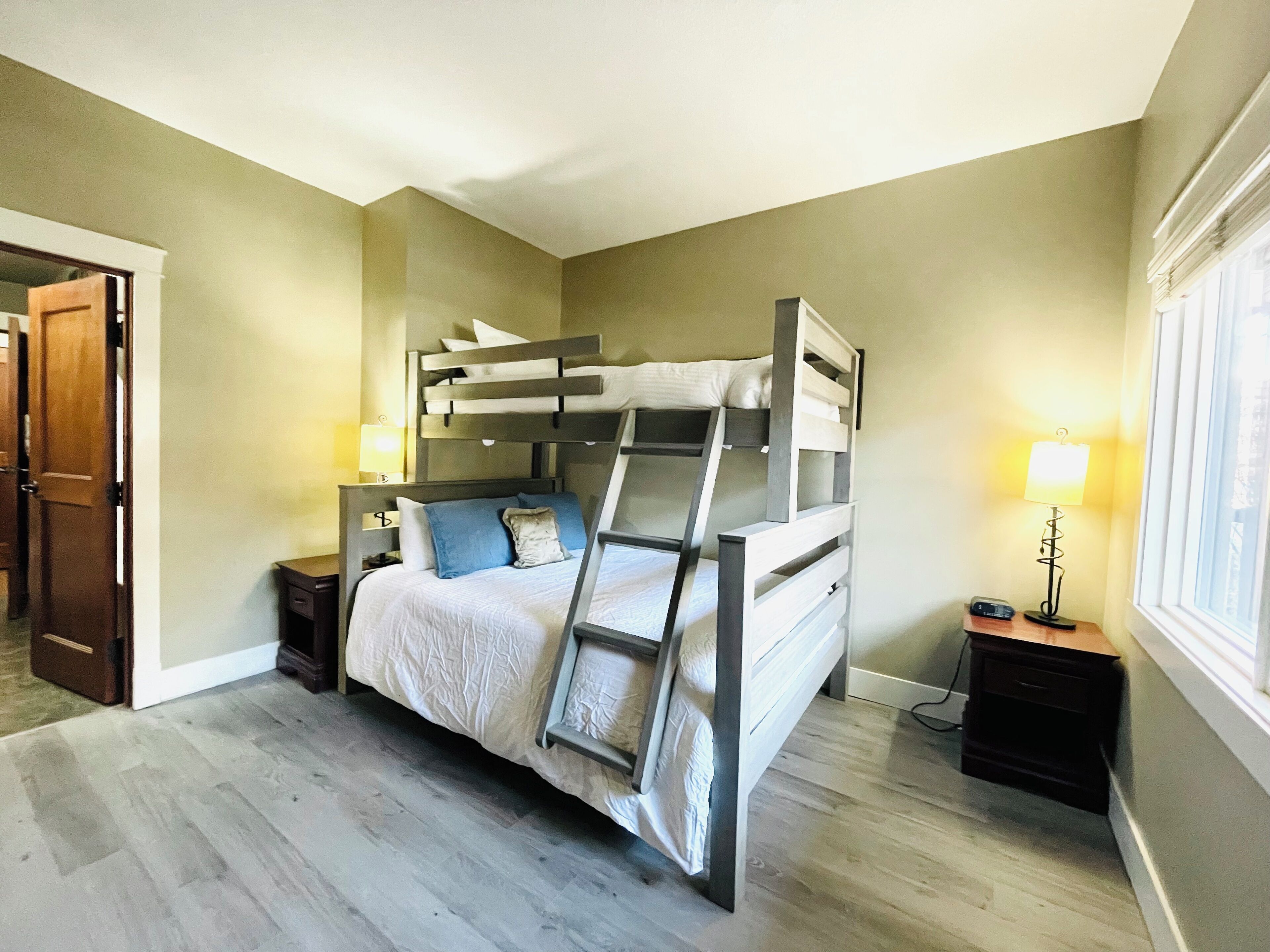 Un lit superposé dans une chambre avec un plancher en bois, une table de chevet avec une lampe et une fenêtre avec des stores.