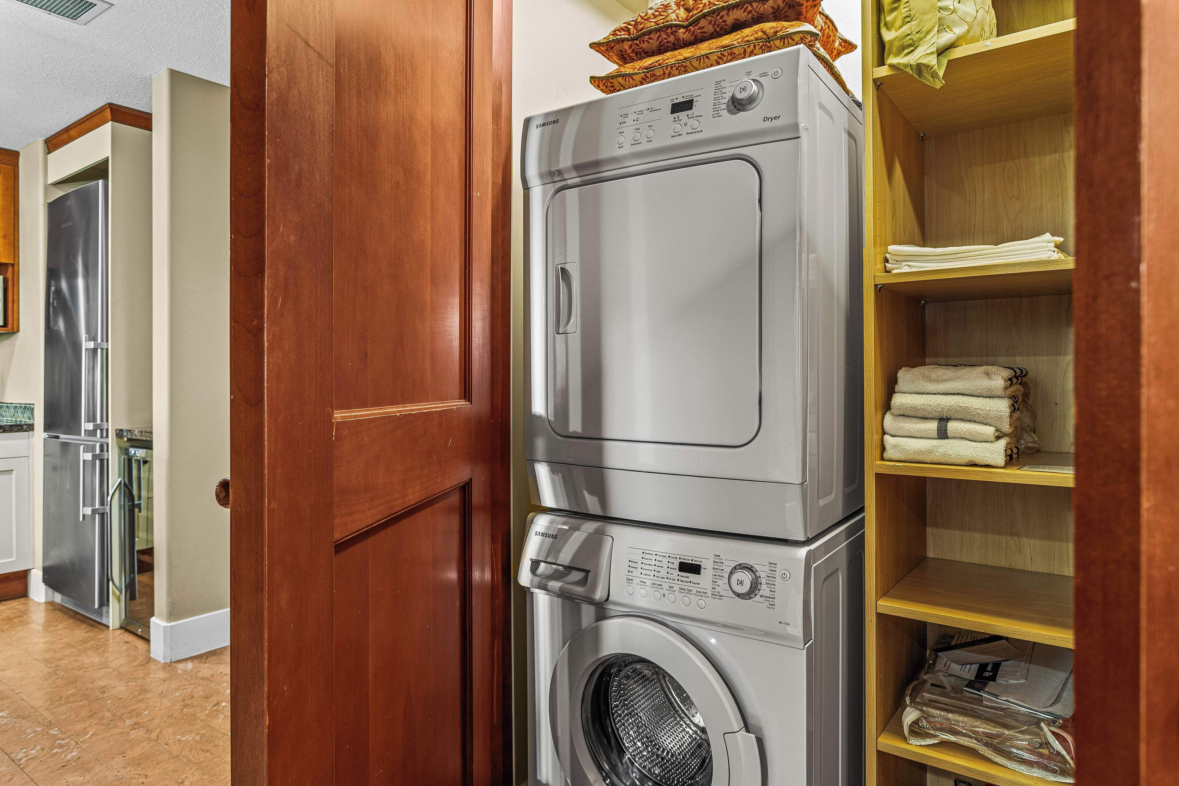 A laundry room with a stacked washer and dryer, wooden shelving with folded towels, and a stainless steel refrigerator.