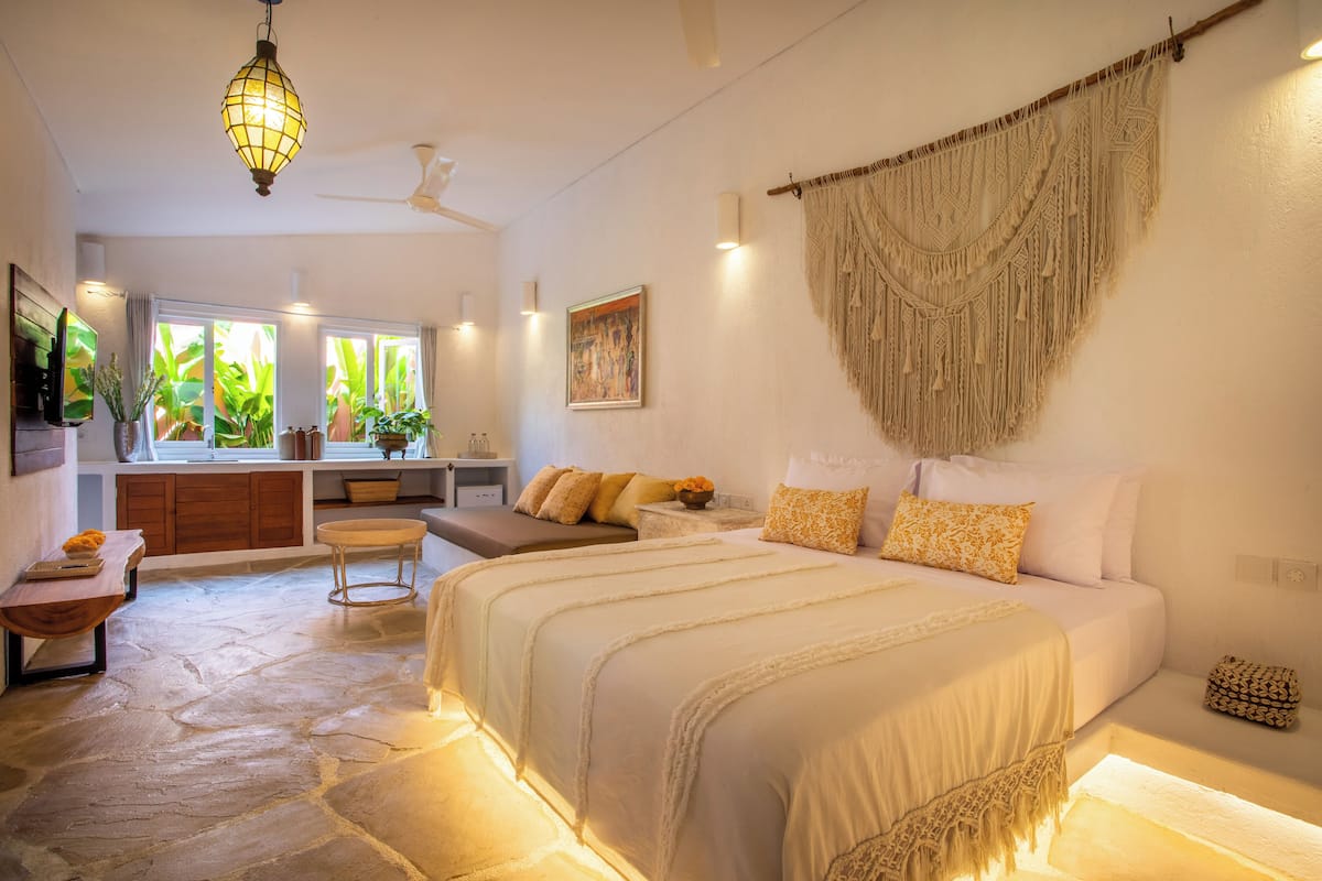A spacious bedroom with a large bed, a sitting area with a bench, a stone floor, and a view of greenery through large windows.