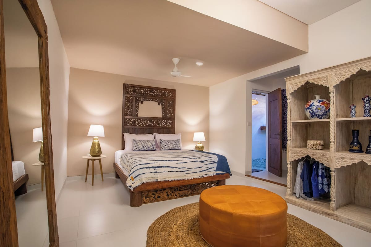 A bedroom with a bed, two bedside lamps, a round ottoman, a decorative mirror, and a wardrobe with shelves.