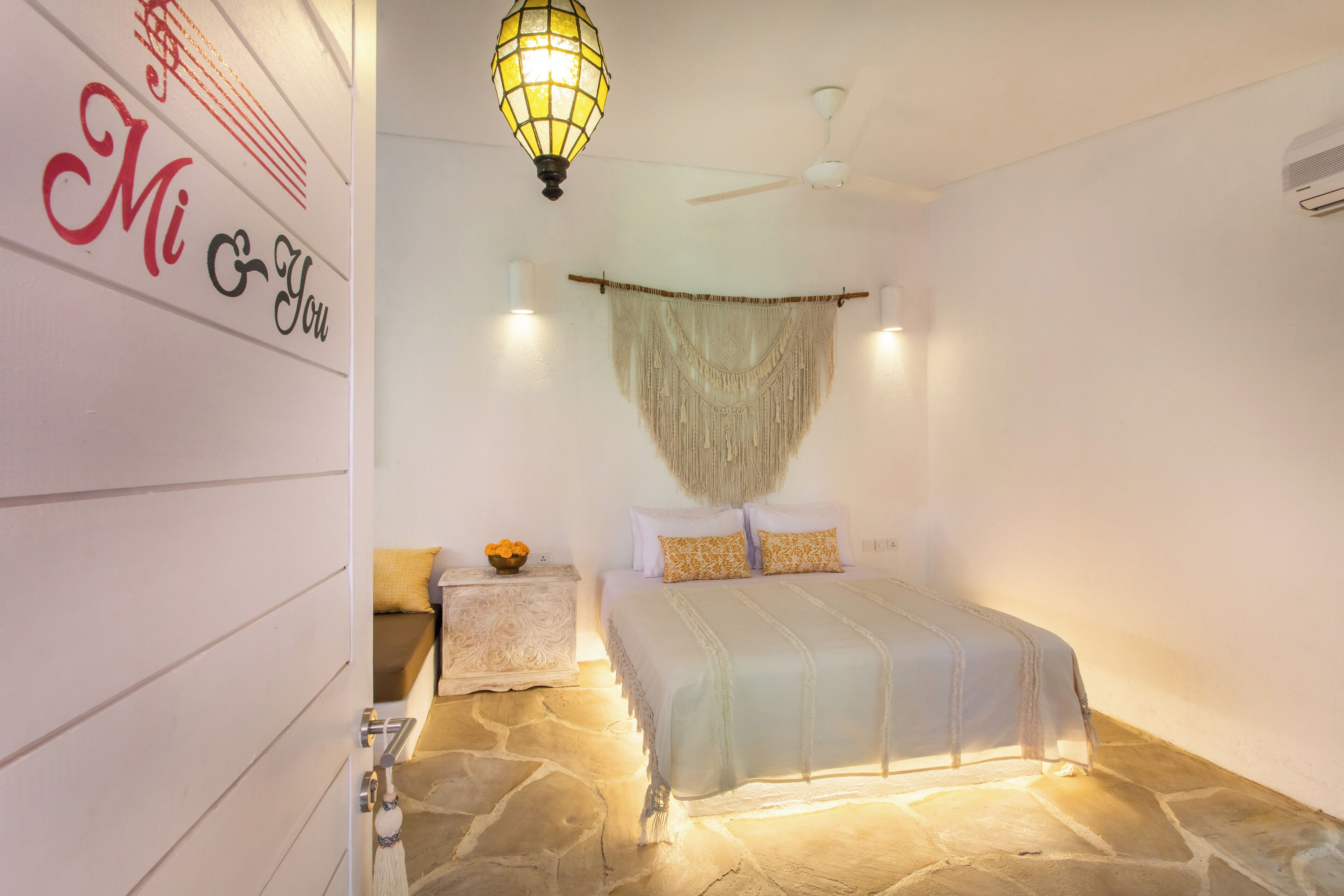 A bedroom with a stone floor, a bed with white and yellow bedding, a wall-mounted macrame piece, a ceiling fan, and a wall with 'Mi & You' decor.