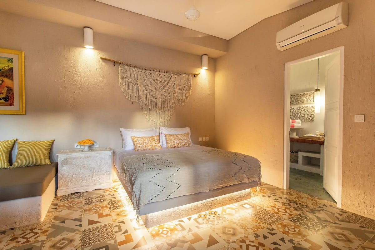 A bedroom with a patterned floor, a bed with a geometric throw, a nightstand with a vase, and a wall-mounted air conditioner.