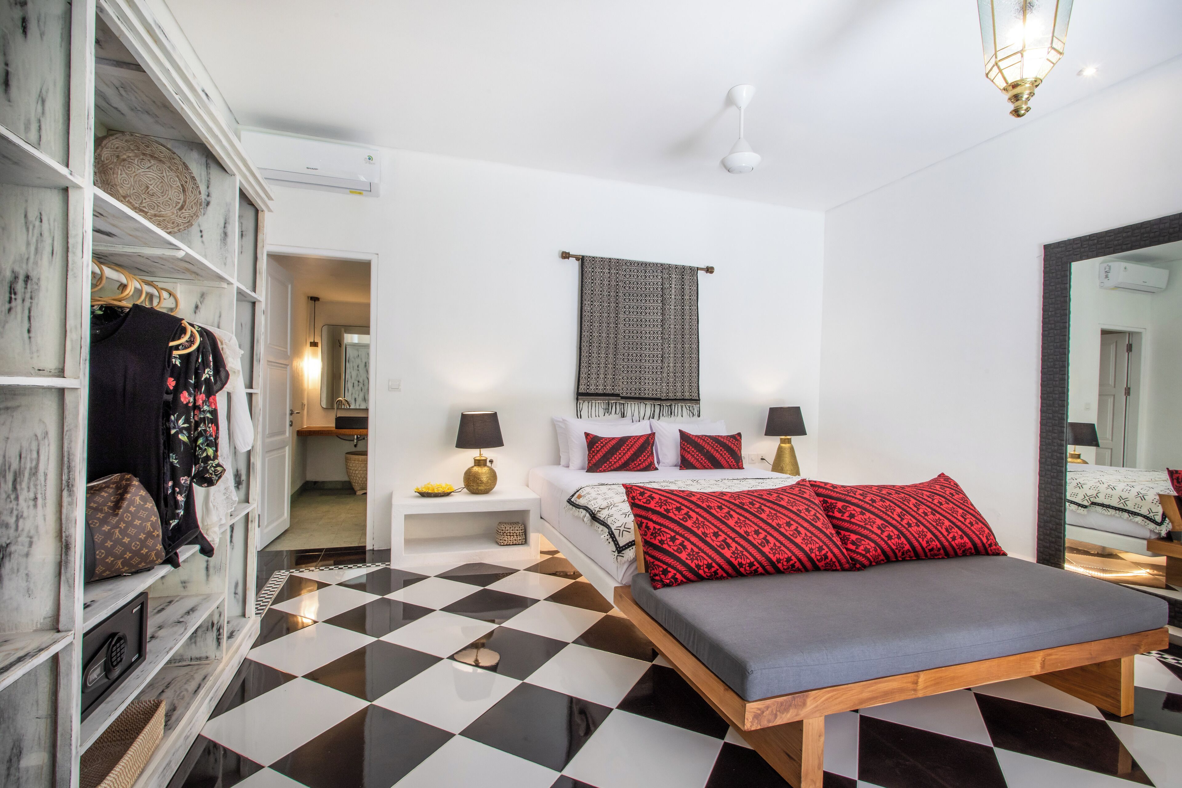 A modern bedroom with a checkered floor, a bed with red and black pillows, a wardrobe with hanging clothes, and a bedside table with a lamp.
