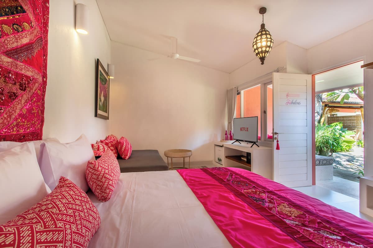 A hotel room with a bed, a desk with a computer, a TV, and a door leading to a balcony with plants.