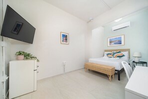 Un dormitorio con cama, televisor montado en la pared, un refrigerador pequeño y una mesita de noche con lámpara.