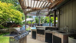 An outdoor kitchen with a grill, sink, and storage cabinets, under a wooden pergola. - Luxury Retreat • Pool, Hot Tub • Cigar Lounge (Santa Rosa)