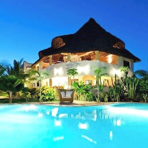 Uma casa grande com telhado de palha, piscina e vegetação tropical exuberante.