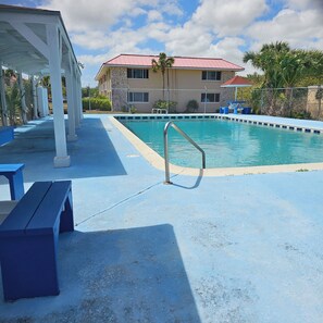 A swimming pool with a metal ladder, blue benches, and a covered area with a building in the background. - Centrally located 2-bed unit w/ washer/dryer, Wi-Fi, pool, and nearby dining. (Freeport)