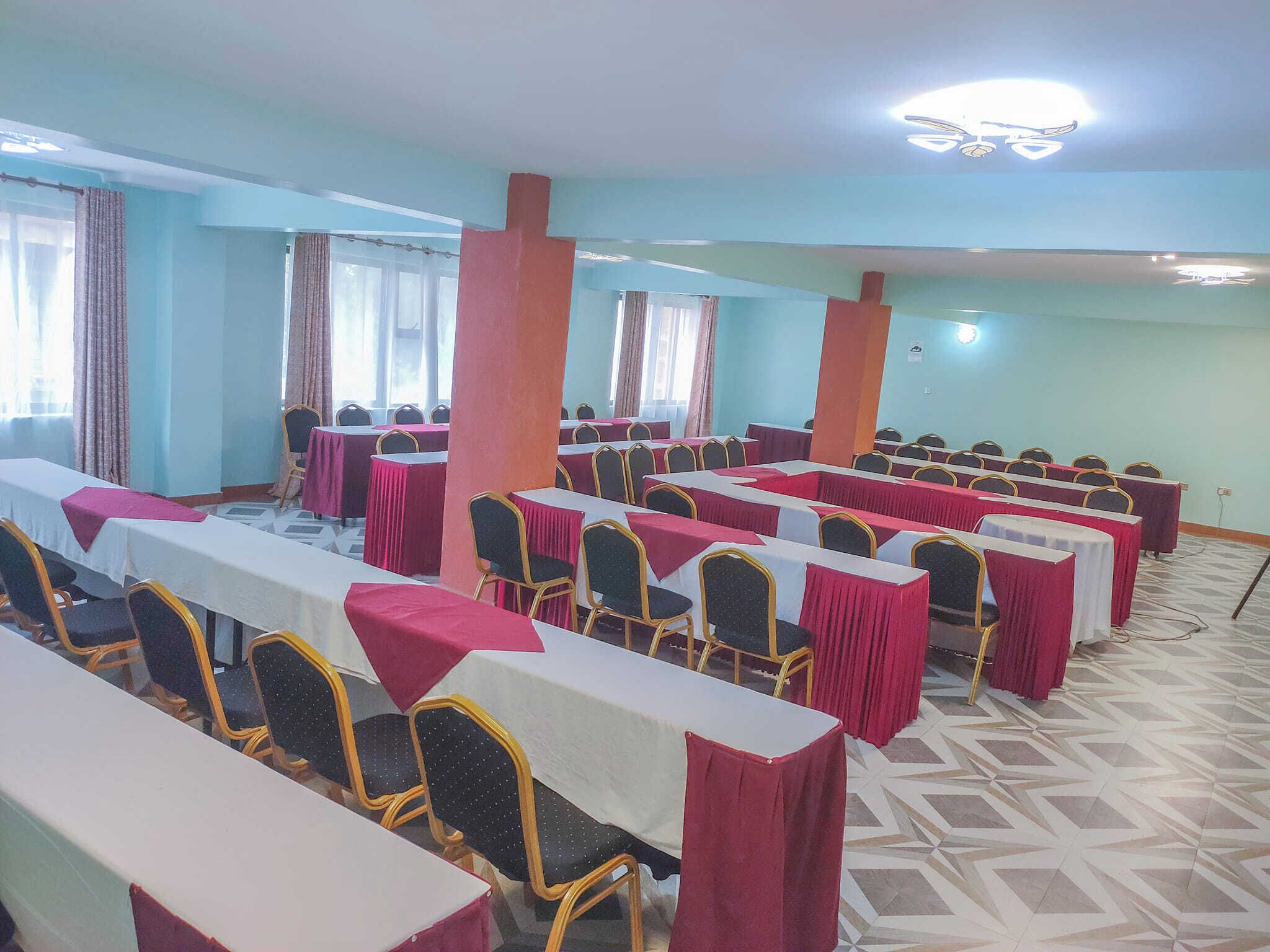 Une salle de conférence avec des rangées de tables et de chaises, de grandes fenêtres avec des rideaux et un sol à motifs.