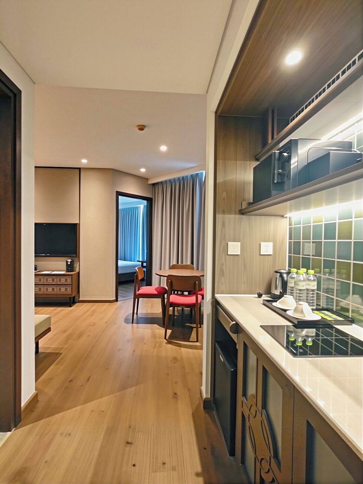 A compact hotel room with a kitchenette, wooden flooring, a dining area with chairs, and a view through a door.
