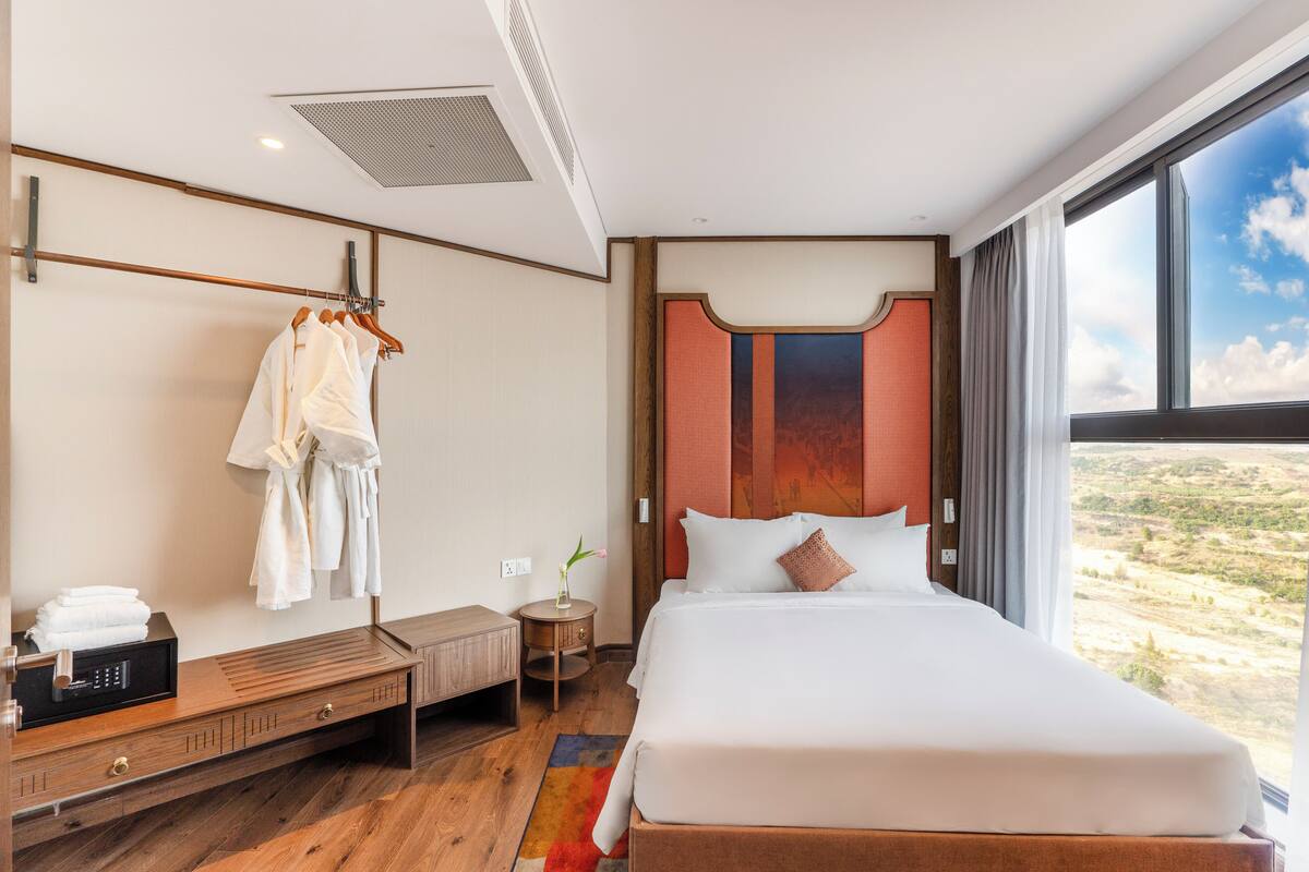 Une chambre d’hôtel avec un grand lit, une table de chevet en bois, une petite table ronde et une vue sur le désert par une grande fenêtre.