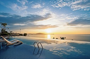 Ein Fernblick vom Infinity-Pool über den Ozean bei Sonnenuntergang, mit Liegestühlen und Palmen.