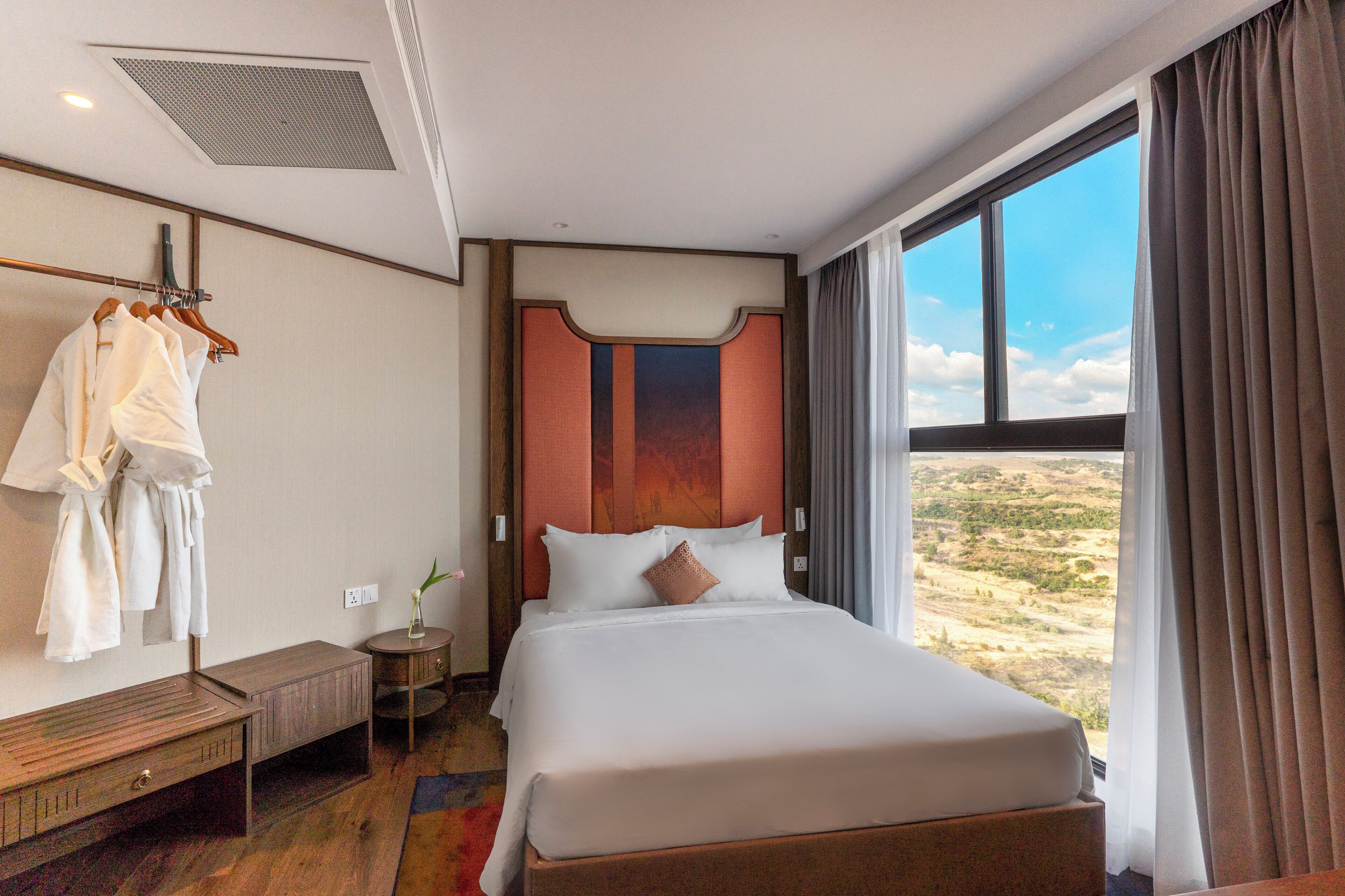 Ein Hotelzimmer mit einem großen Bett, einer hölzernen Bank, einem Bademantelständer und Blick auf die Wüste durch das Fenster.