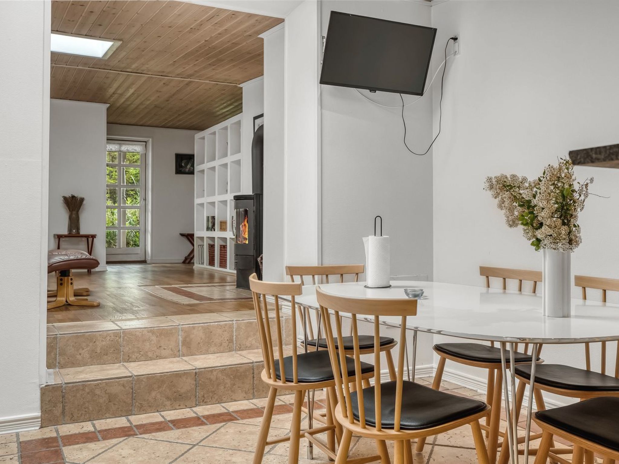 Un espace repas moderne avec une table blanche, des chaises en bois et un téléviseur à écran plat fixé au mur.