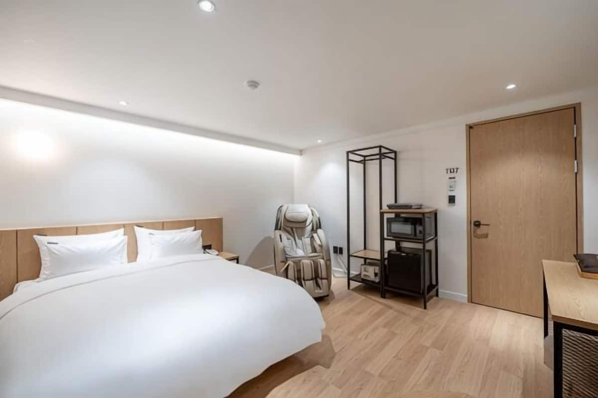 A modern hotel room with a large bed, a massage chair, a small table, and a door marked '107'.