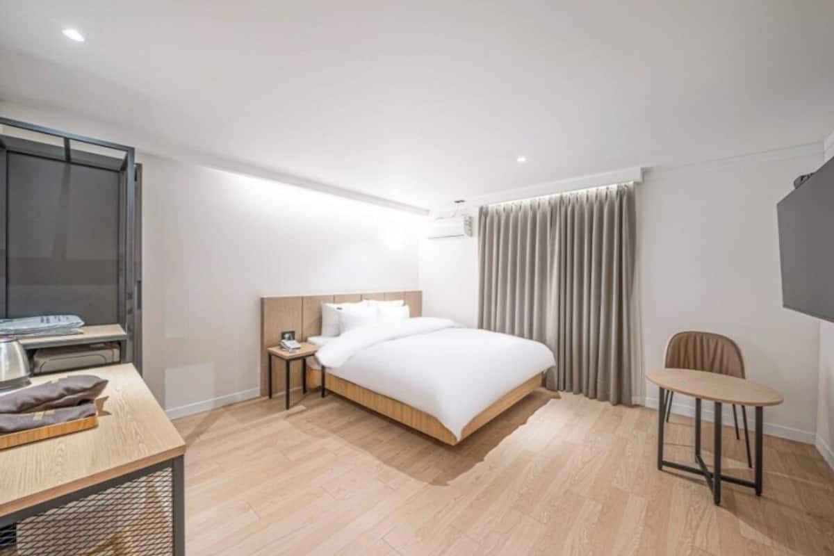 A modern hotel room with a wooden bed, a desk with a kettle, a chair, and a large window with curtains.