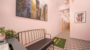 Un hall d’hôtel avec un banc, un comptoir d’accueil, un tapis vert sur lequel est inscrit le nom « HÔTEL LUZ », et un grand tableau représentant une scène de rue.