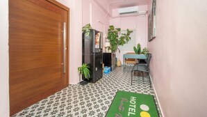 Un couloir étroit avec une porte en bois, un réfrigérateur, une petite table avec des chaises et un tapis vert orné du nom de l’hôtel.