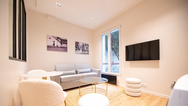 Un salon moderne avec un canapé gris, des fauteuils blancs, une table basse en verre et une grande fenêtre donnant sur un paysage arboré.