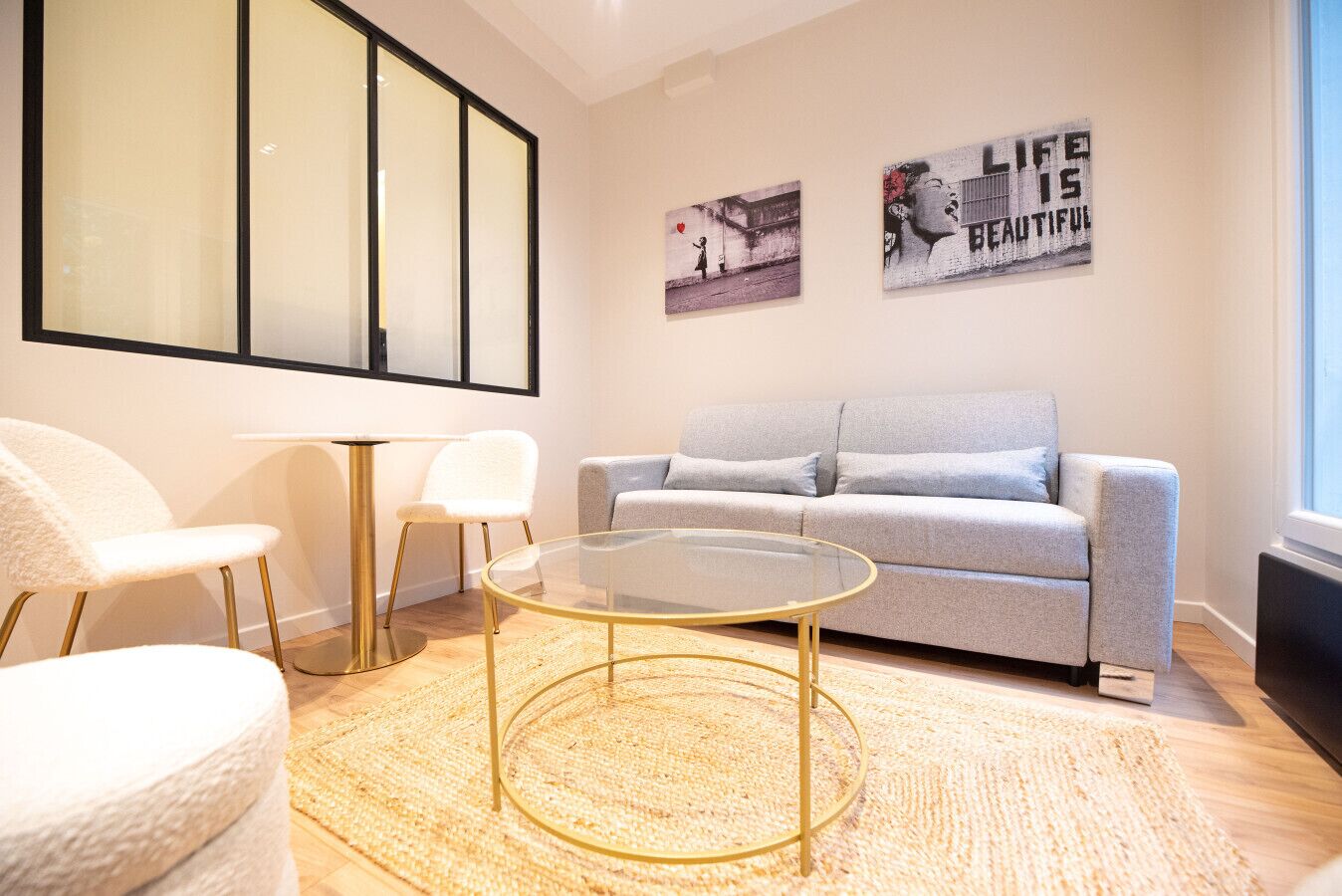 A modern living room with a grey sofa, a round glass coffee table, and a white armchair. There are two framed pictures on the wall, one depicting a cityscape and the other a quote.