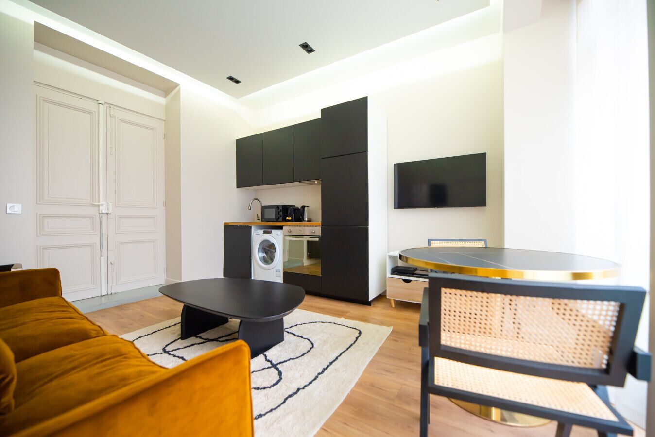 Un espace de vie compact comprenant un canapé, une table à manger, un coin cuisine équipé d’appareils électroménagers et un téléviseur fixé au mur.