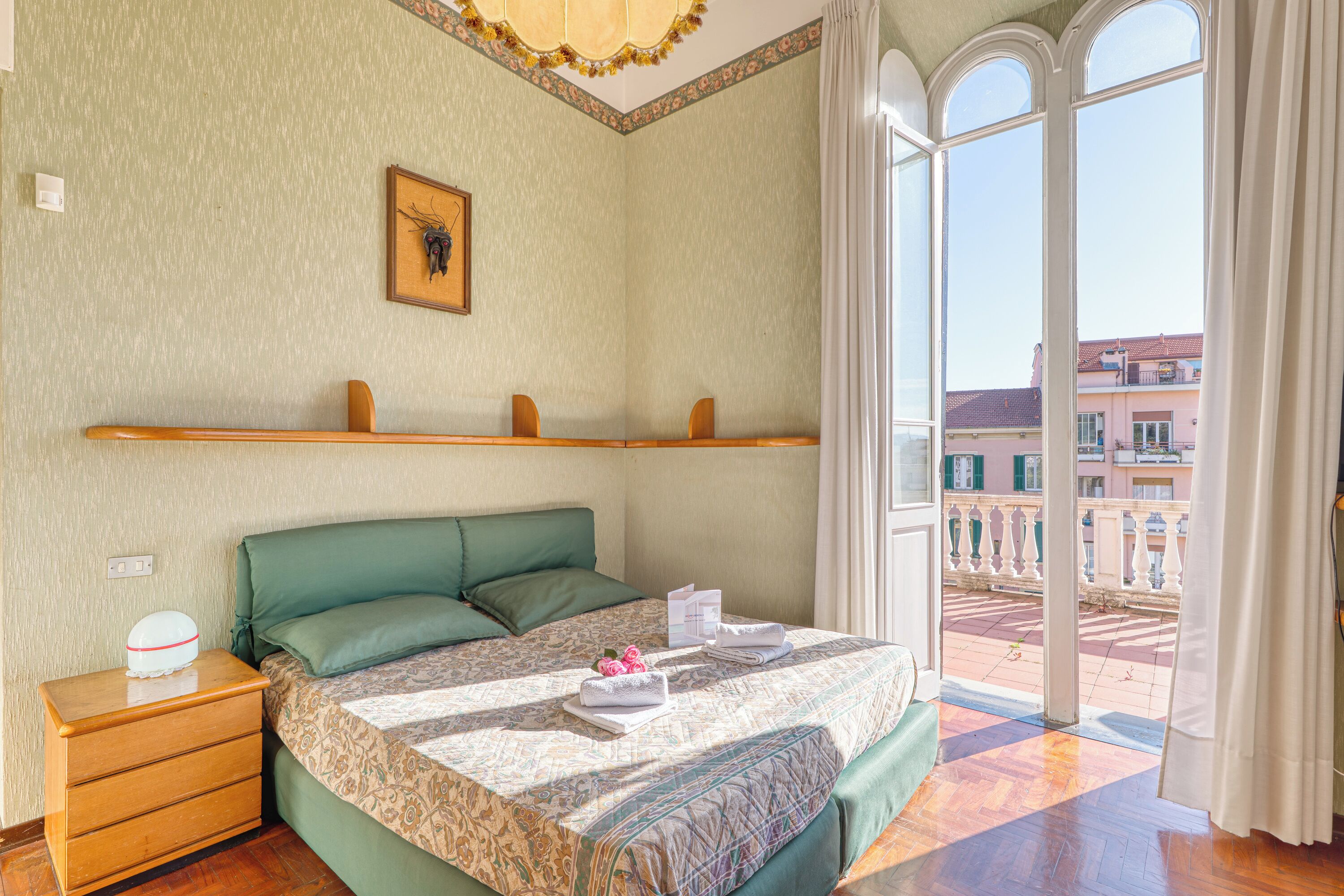 Una habitación de hotel con cama, mesita de noche, ventana con vistas a edificios e iluminación decorativa en el techo.