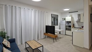 Ein modernes Wohnzimmer mit einem blauen Sofa, einem Couchtisch und einem Fernseher. Es gibt eine Küchenzeile mit Herd, Mikrowelle und Schränken.