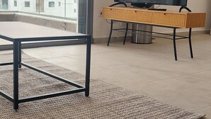 Ein modernes Wohnzimmer mit großem Fenster, einem Flachbildfernseher auf einem Holzständer, einem Couchtisch und einem Teppich mit Muster.