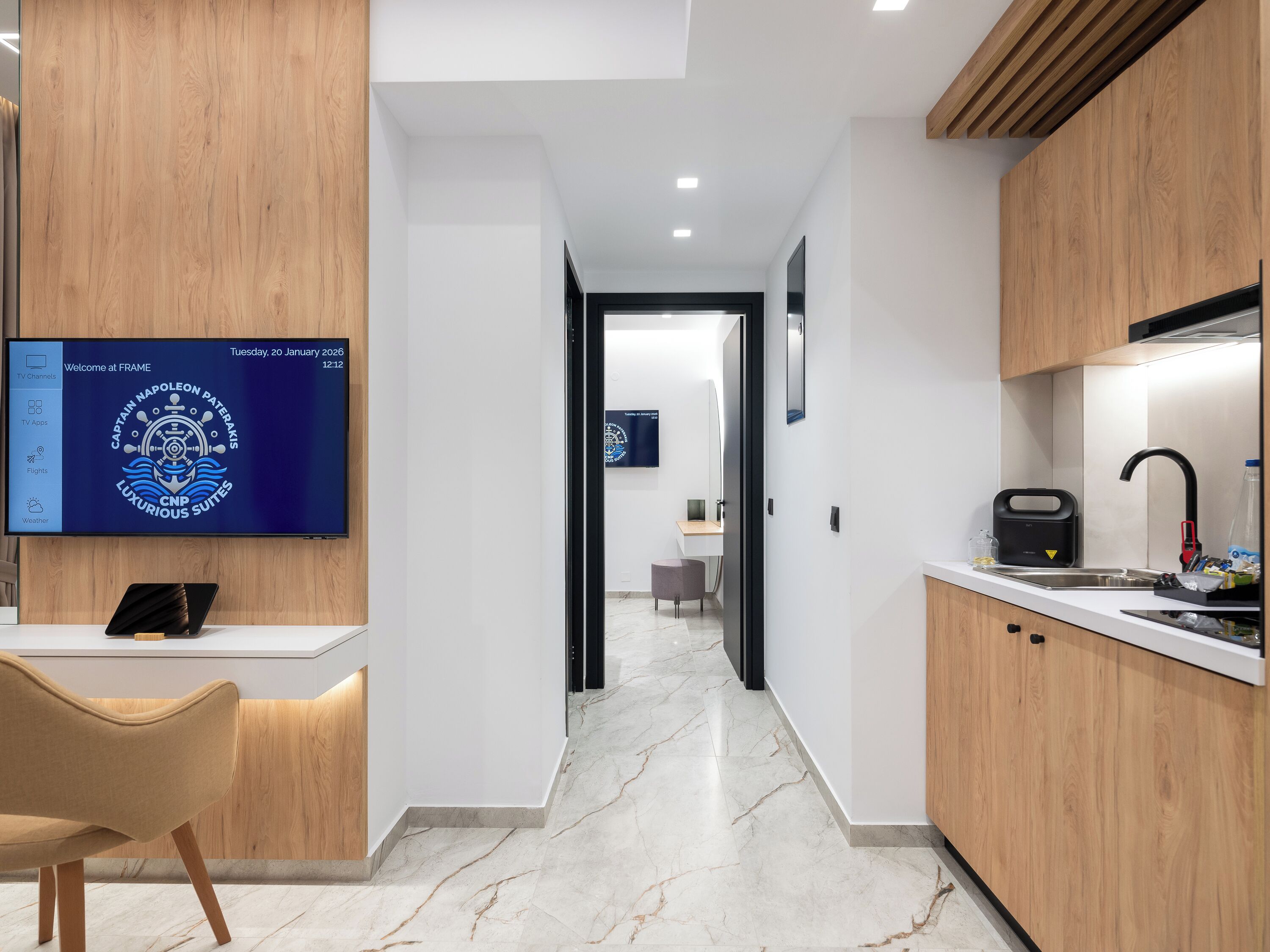 A modern hotel room with a wooden accent wall, a flat-screen TV displaying a welcome screen, a marble floor, and a kitchen area with a sink and countertop.