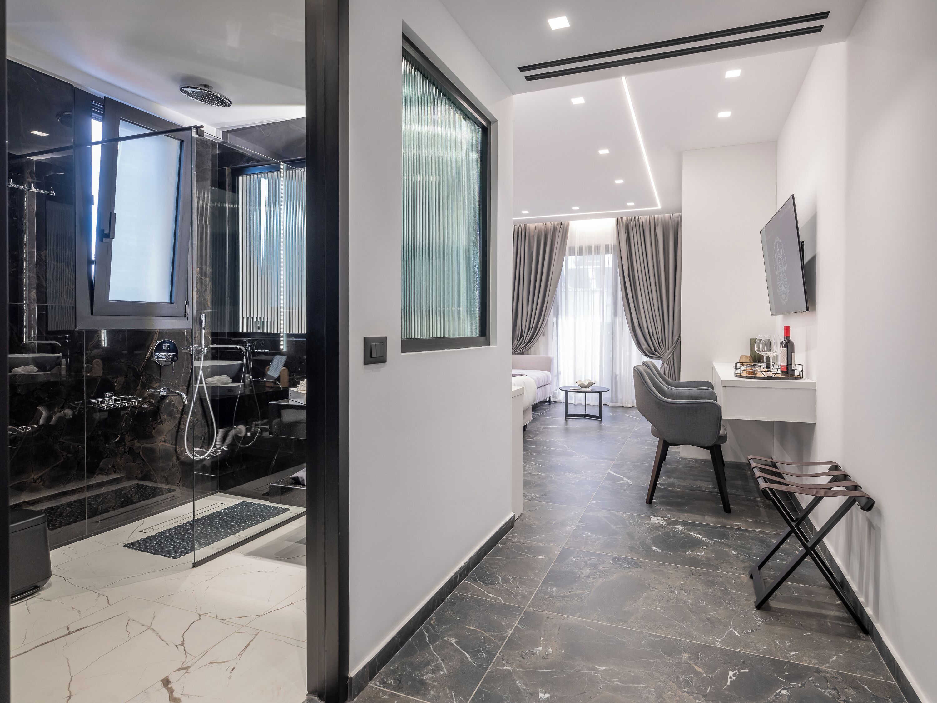 A modern hotel room with a glass-enclosed shower, a bed with a view, a desk with a chair, and a TV mounted on the wall.